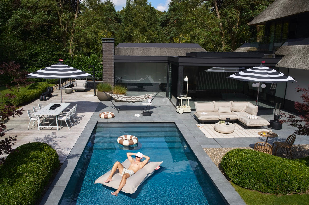 Man relaxing on a beige Floatzac floating bean bag in a backyard pool surrounded by outdoor furniture, garden, and shaded seating areas