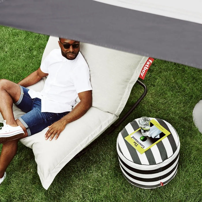 A man relaxes on a soft garden lounger while a black-and-white striped Fatboy Point Outdoor Pouf beside him holds a drink and a book, styled as part of a casual seating area by Desert River Shop.