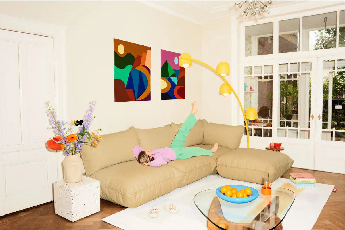  A person relaxes on a beige Fatboy Sumo Weave sectional sofa in a sunlit living room, complemented by a vibrant yellow Big Lebow floor lamp and colorful abstract wall art.