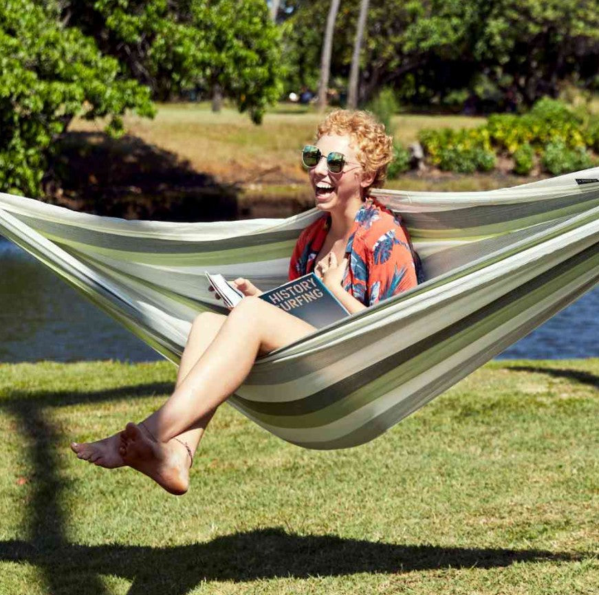 Smiling woman relaxes in the Brisa KingSize Hammock by the lake while holding a book.